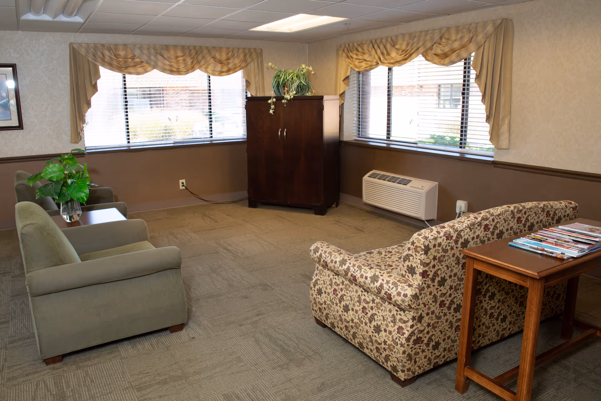 A small lounge with upholstered chairs and a patterned sofa, a side table with magazines, a potted plant, and large windows with valances.