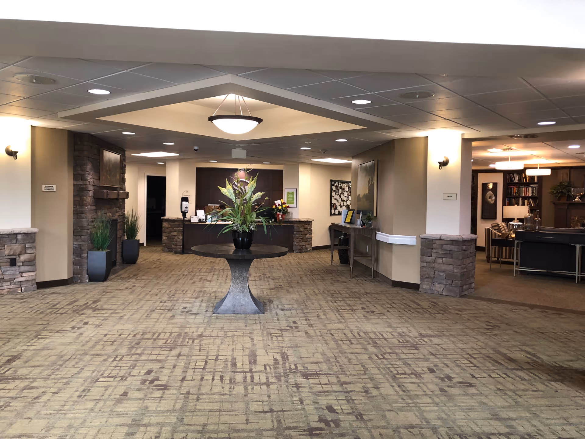 Interior view of a senior living facility lobby with a round table holding a floral arrangement in the center. The space features stone accents on the walls, a reception desk in the background, potted plants, and a seating area with chairs and bookshelves to the right.