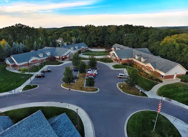 Aerial view of a senior living facility with multiple single-story buildings arranged around a circular driveway with parked cars, surrounded by green lawns and trees under a partly cloudy sky.