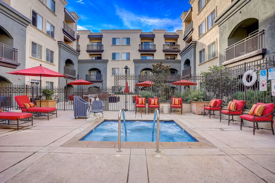 Outdoor courtyard area of a senior living facility with a small rectangular pool or hot tub in the center, surrounded by red cushioned lounge chairs and umbrellas. The area is enclosed by a metal fence and is flanked by multi-story residential buildings under a blue sky with some clouds.