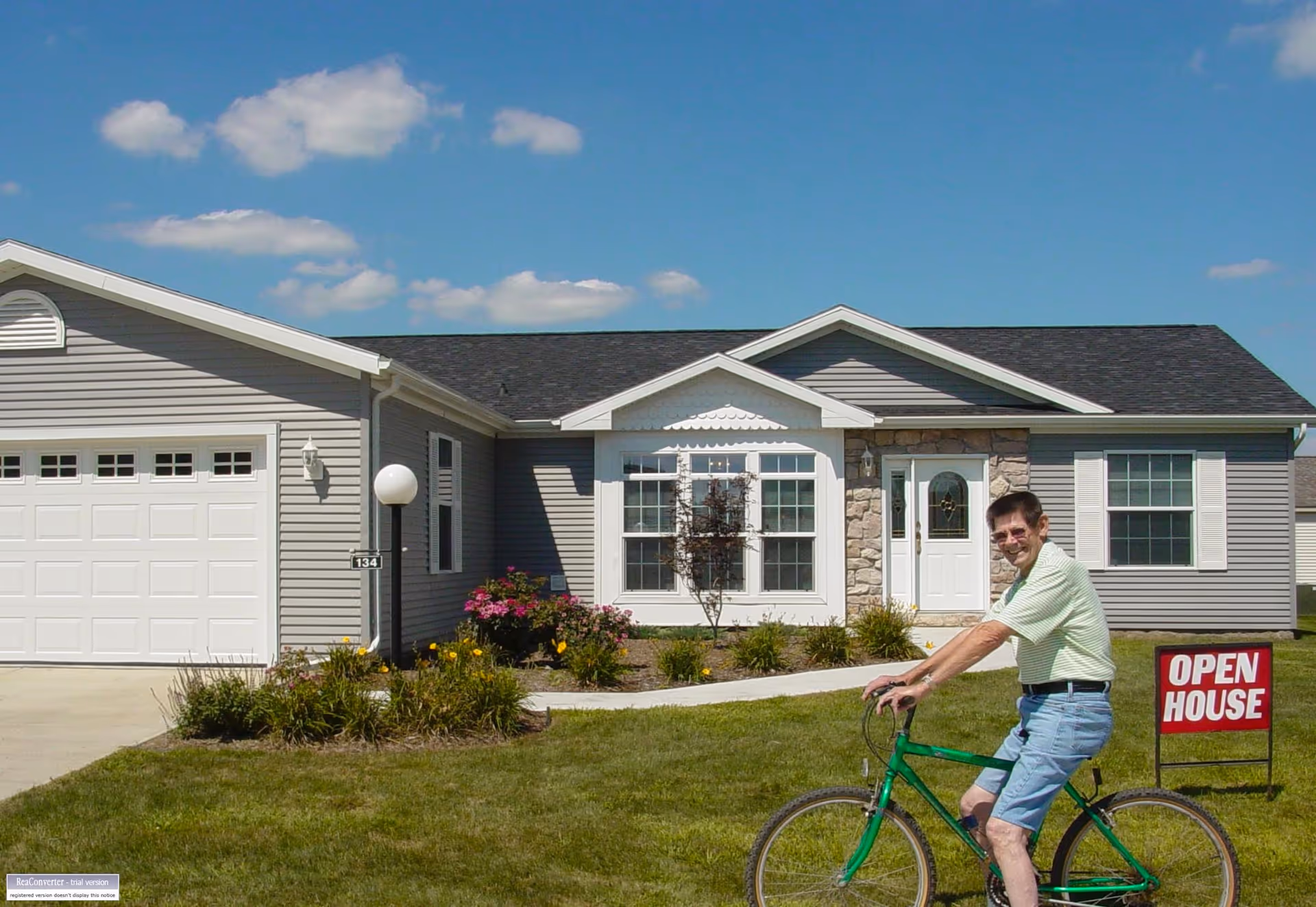 A man wearing a light green striped shirt and blue shorts is sitting on a green bicycle on the lawn in front of a single-story house with gray siding, white trim, and a stone accent around the front door. There is an 'OPEN HOUSE' sign on the grass near the sidewalk leading to the front door. The sky is blue with some scattered clouds.