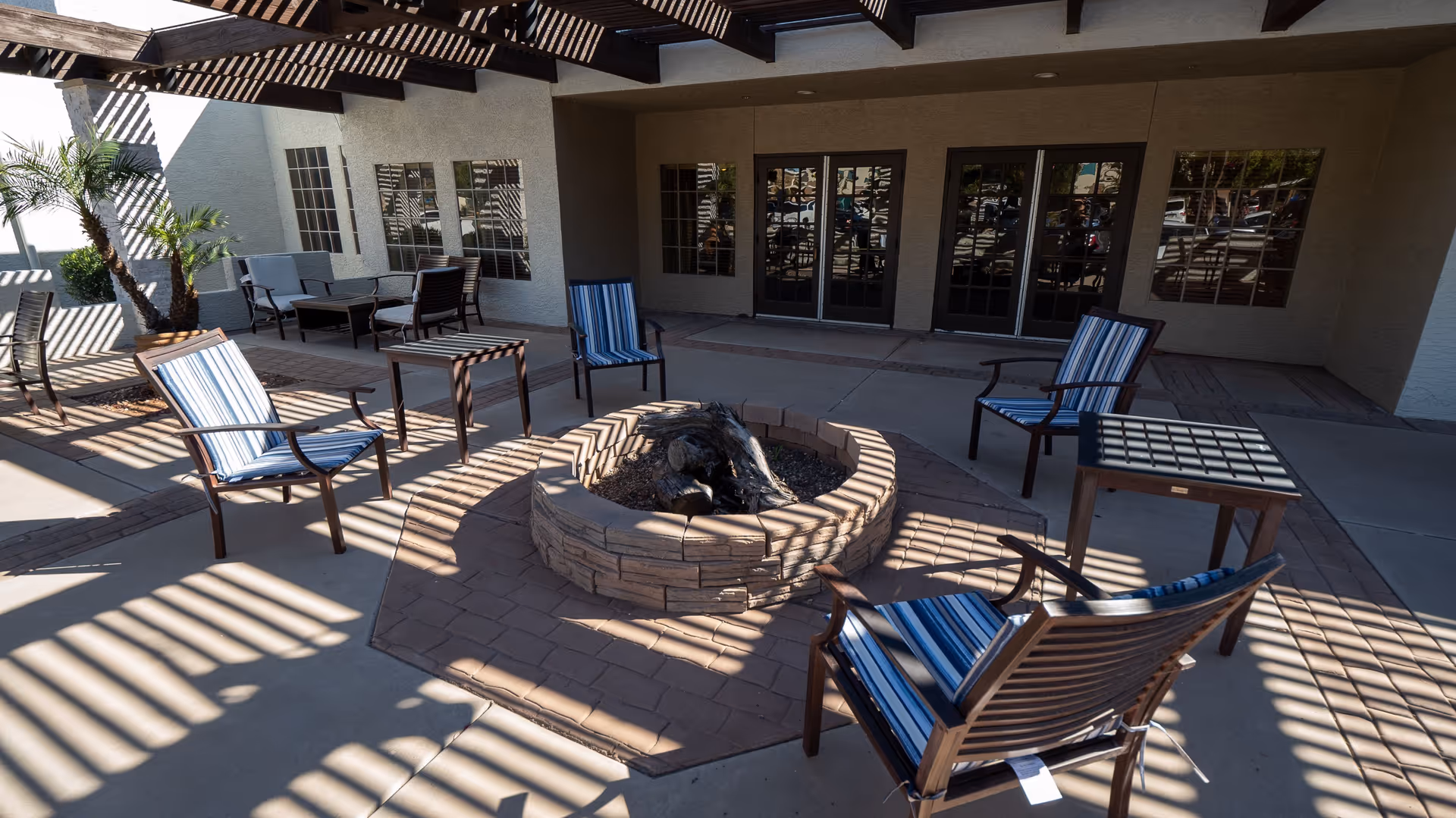 Outdoor patio area with a circular stone fire pit surrounded by six chairs with blue and white striped cushions. The patio is shaded by a pergola casting striped shadows on the ground and furniture. There are several windows and glass doors on the building wall behind the seating area, and some potted plants are visible on the left side.