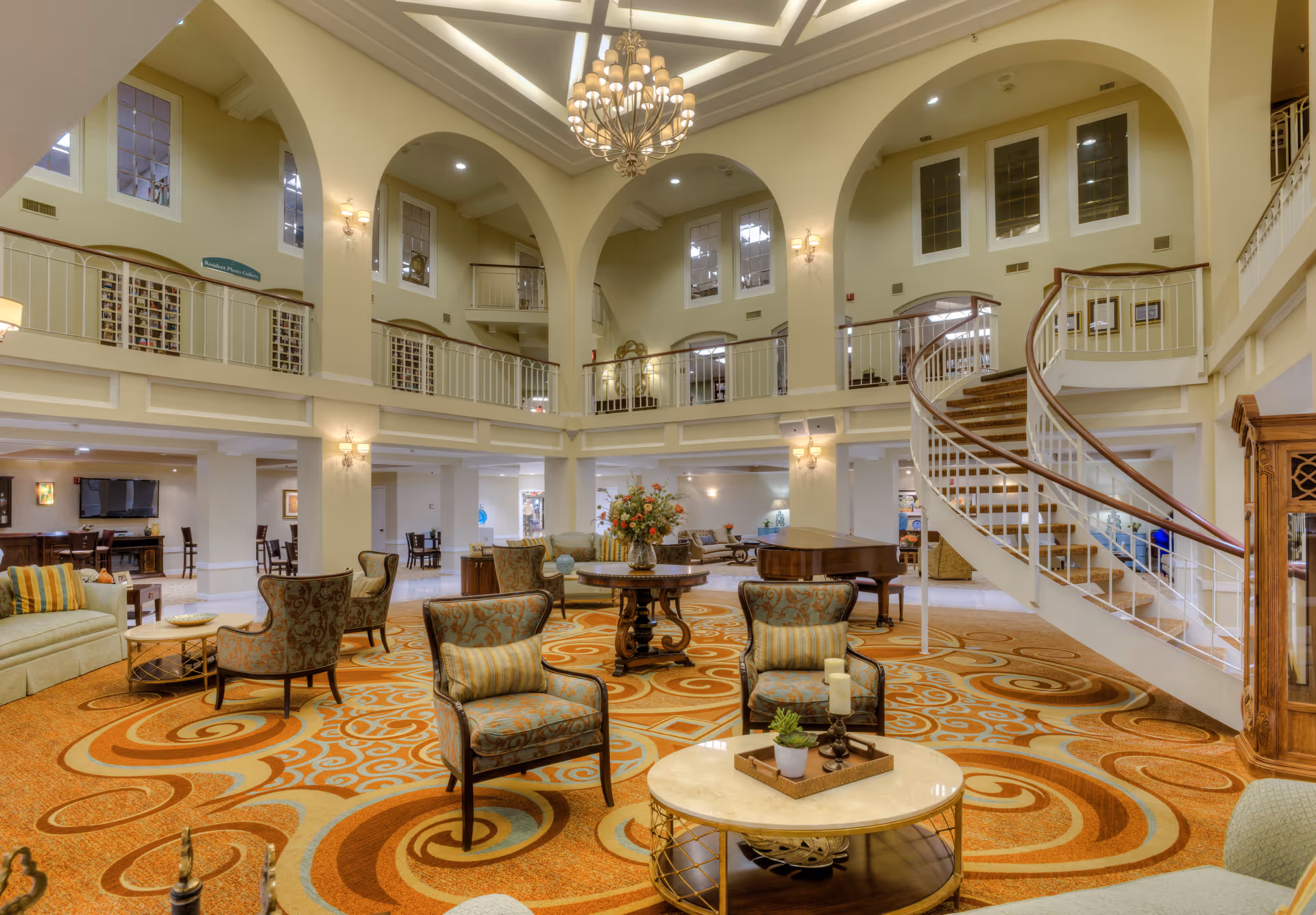 Spacious and elegant senior living facility common area with high ceilings, large arches, and a decorative chandelier. The room features a patterned orange and beige carpet, multiple seating arrangements with upholstered chairs and sofas, a round table with a floral centerpiece, and a grand piano near a curved staircase leading to an upper balcony.
