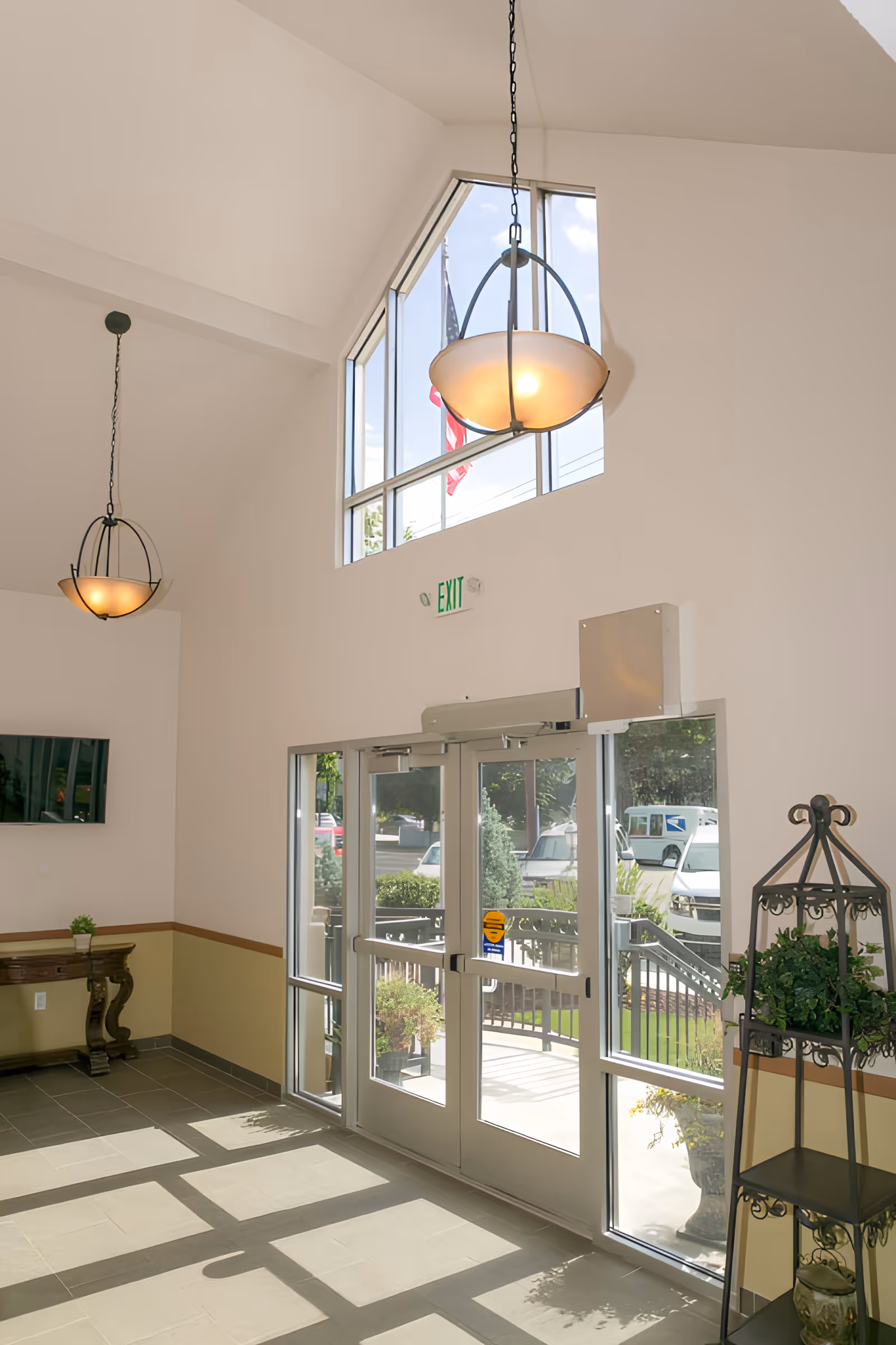 Interior view of a bright entrance area with large glass double doors and tall windows above letting in natural light. Two hanging pendant lights are visible from the high ceiling. There is a small wooden table with a plant on it to the left and a decorative metal plant stand with greenery on the right. Outside, a flagpole with an American flag is visible.