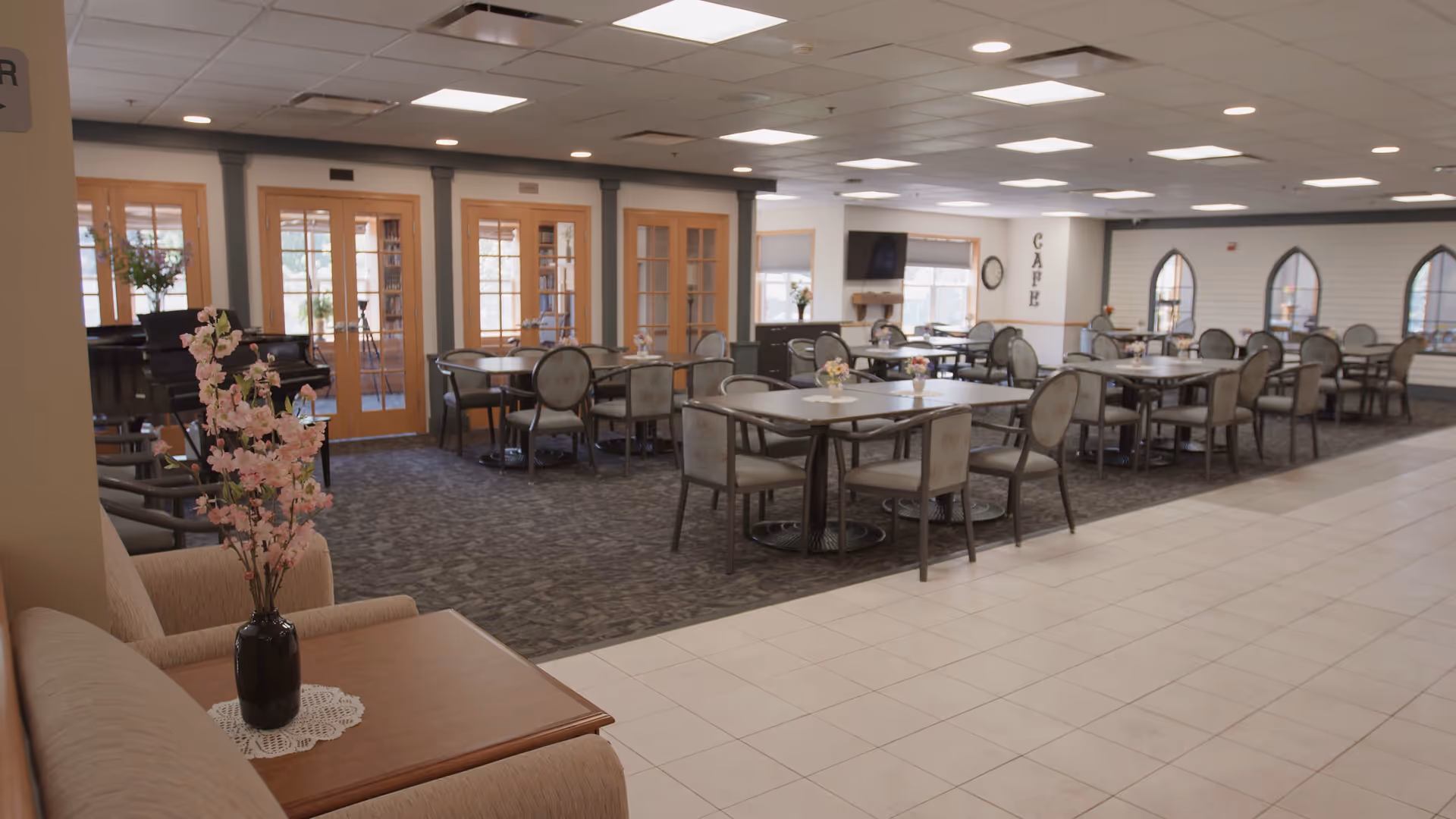 A spacious dining area in a senior living facility with multiple tables and chairs arranged neatly. The room features a carpeted section with tables and chairs, a tiled floor area, a sofa with a wooden side table holding a vase of pink flowers, and a piano near wooden double doors with glass panels. The word 'CAFE' is displayed vertically on a column near windows with natural light coming in.