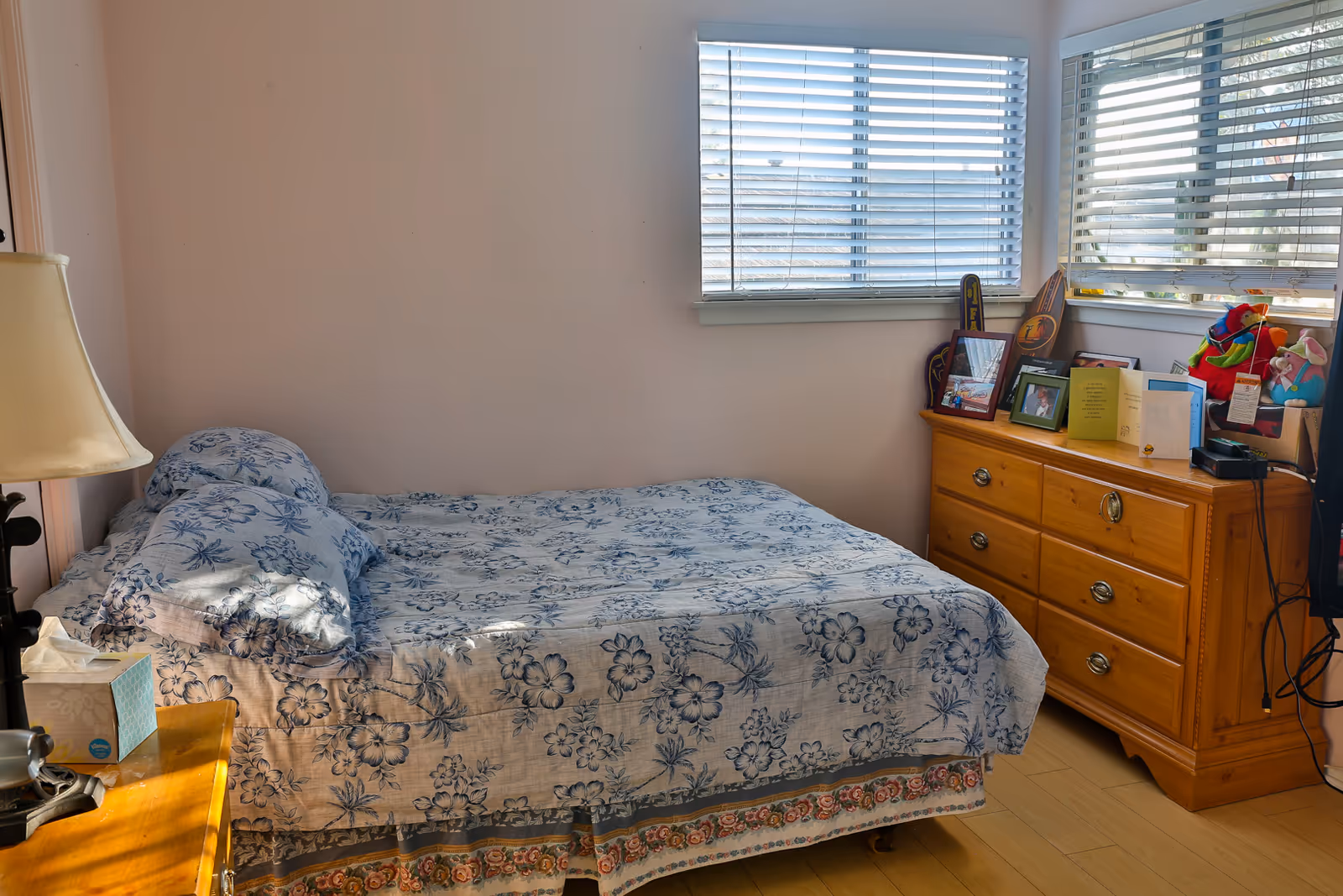 Small bedroom with a floral bedspread, wooden nightstand and dresser with photos and cards, and two windows with blinds.