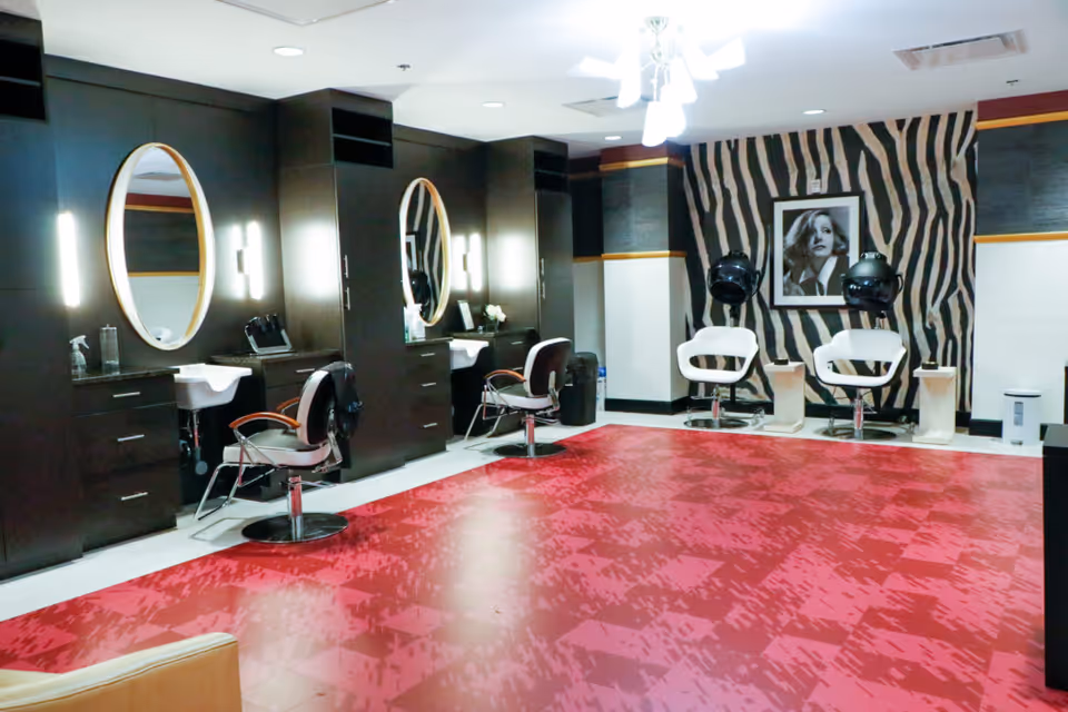 Interior view of a salon area with two styling stations featuring round mirrors, chairs, and sinks on the left side. On the right side, there are two white chairs with black hair dryers above them, set against a zebra-striped wall with a black and white portrait of a woman. The floor has a red patterned design.