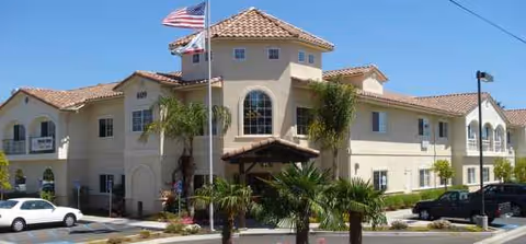 Tan two-story senior living facility with a tiled roof, flagpoles, and palm trees.