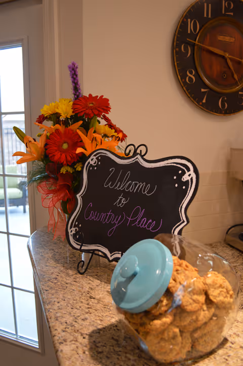 A kitchen countertop with a bouquet of colorful flowers, a decorative chalkboard sign that says 'Welcome to Country Place,' and a glass jar filled with cookies with a blue lid. A large wall clock is visible on the wall above the counter, and a glass door leading outside is in the background.