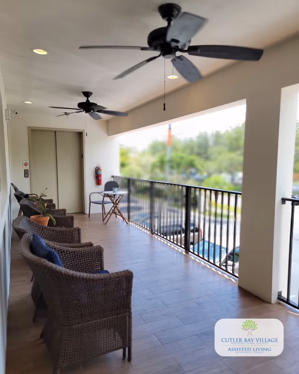 A covered outdoor balcony area with several wicker chairs arranged along the left side, a small table with two chairs near the railing, ceiling fans, and an elevator door at the far end. The balcony overlooks a parking lot and greenery.
