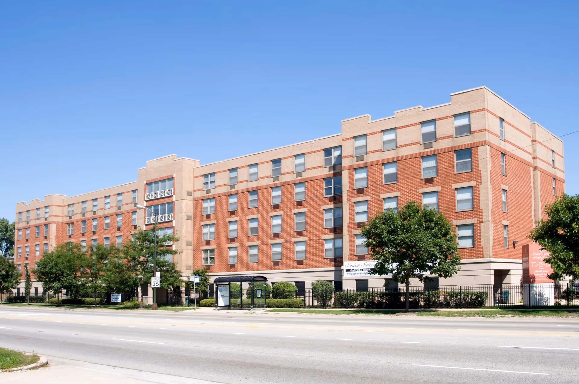 Exterior view of a four-story brick senior living facility building with multiple windows, trees, and a bus stop shelter in front under a clear blue sky.