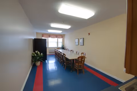 A long rectangular dining room with a large table surrounded by wooden chairs. The floor has a blue and red pattern, and there is a window at the far end with a valance. The walls are light-colored, and there are two framed pictures on one wall. A refrigerator and a plant are visible near the window.