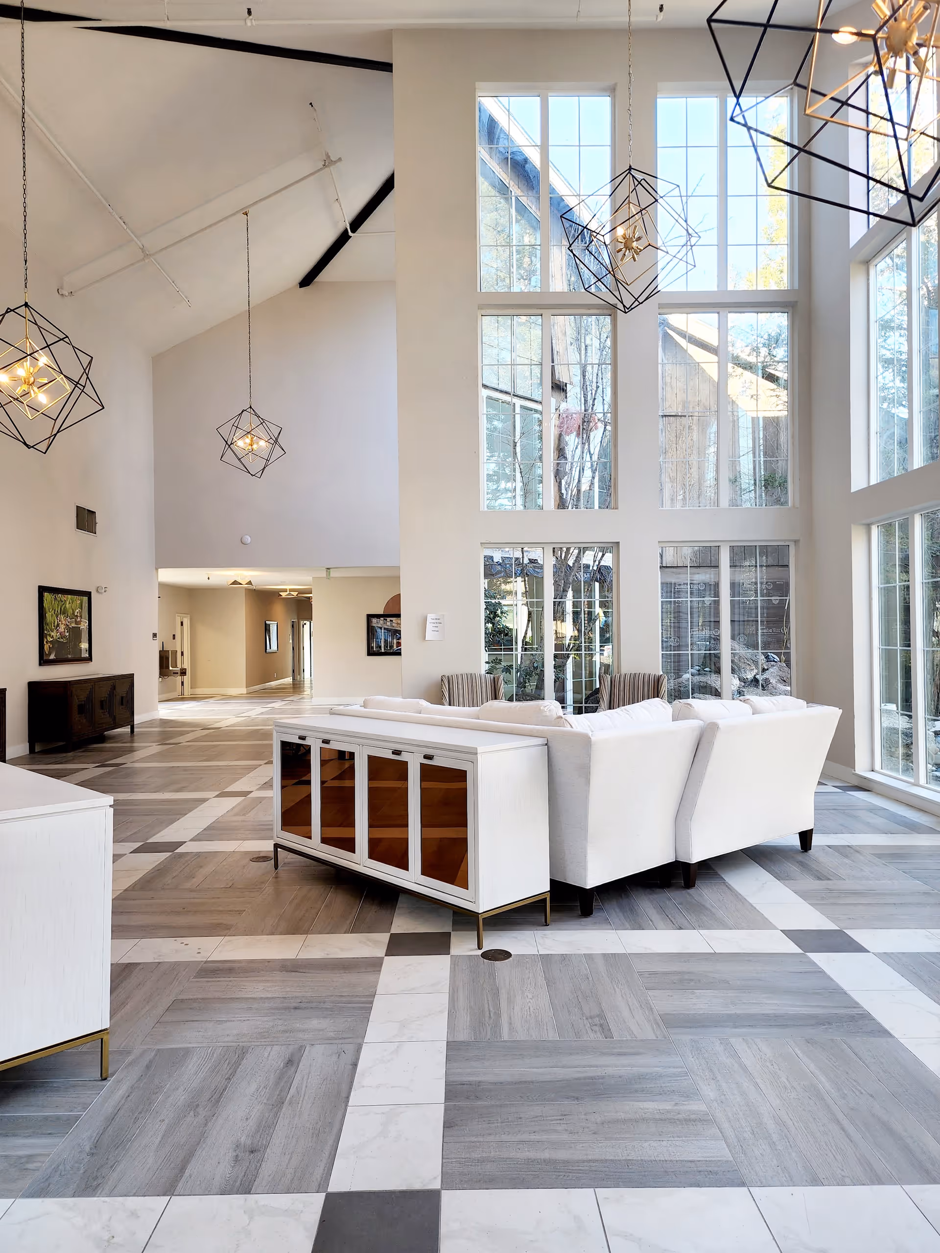 Bright and spacious living area with high vaulted ceilings and large floor-to-ceiling windows allowing natural light to flood the space. The room features modern geometric pendant lights, a white sectional sofa, and a white cabinet with glass doors. The floor has a patterned tile design in shades of gray and white. The overall decor is contemporary and airy.