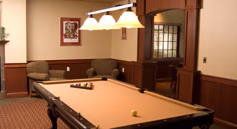 Interior game room with a pool table under pendant lights, upholstered chairs, and wood-paneled walls.