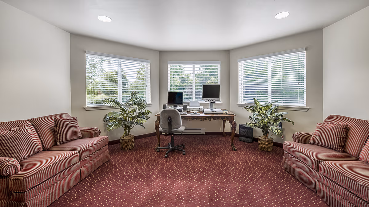 Carpeted common room with two striped sofas facing a desk with a computer beneath three windows, flanked by potted plants.
