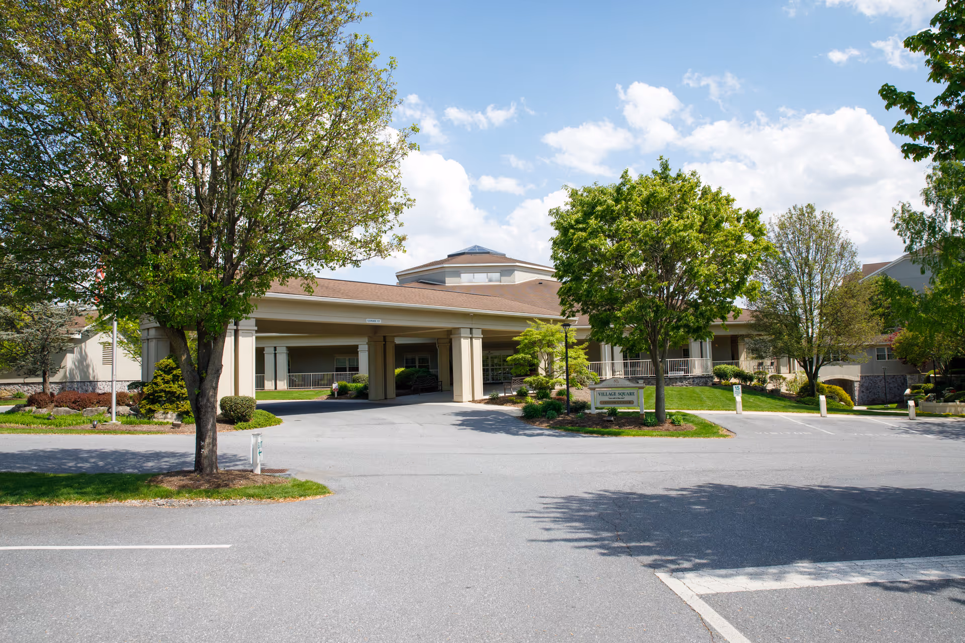 Exterior view of a senior living facility building with a covered entrance, surrounded by green trees and landscaping under a partly cloudy sky.