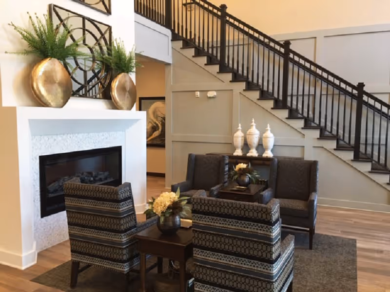 A cozy lobby lounge with patterned armchairs around a coffee table, a fireplace, decorative vases, and a staircase in the background.