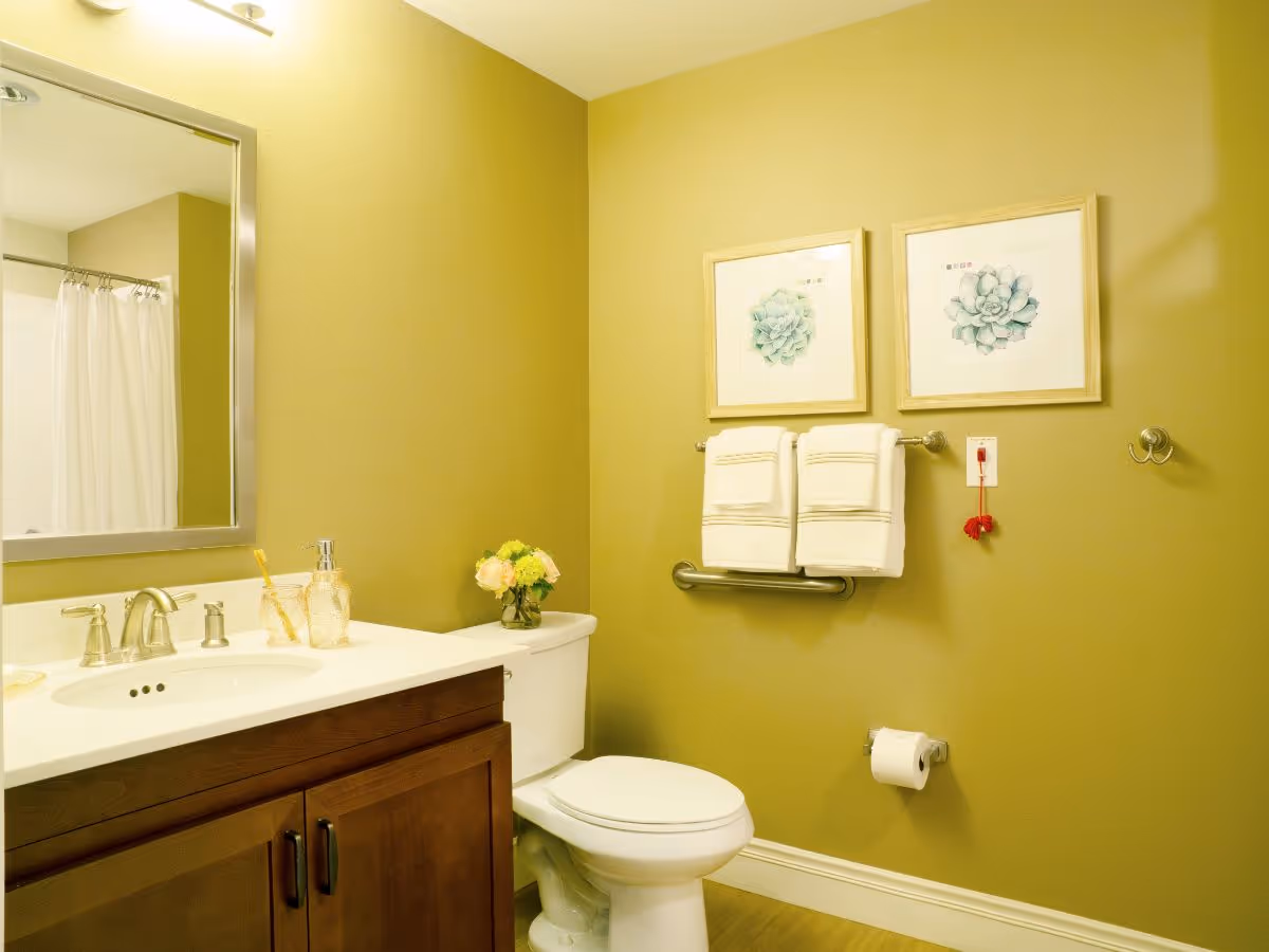 A clean bathroom with a white toilet, a wooden vanity with a white countertop and sink, a large mirror above the sink, two framed succulent artworks on the wall, two neatly folded white towels hanging on a grab bar, a toilet paper holder with a roll, and a small vase with flowers on the toilet tank.