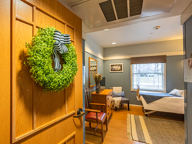 View into a cozy bedroom in a senior living facility with a wooden door decorated with a green wreath and striped bow. Inside the room, there is a bed with white and blue bedding near a window with blinds and a valance. A wooden dresser with a vase of dried flowers, a chair with a blanket, a wall mirror, and framed pictures are also visible. The room has wooden flooring and a striped rug.