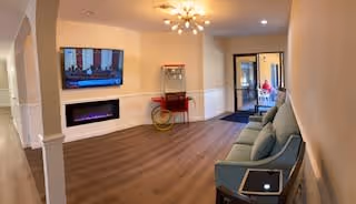 A cozy living room area in a senior living facility with a mounted flat-screen TV above a modern electric fireplace. There is a popcorn machine on a small red cart against the wall, and a light blue sofa with a tablet resting on one arm. The room has wood flooring, warm lighting from a ceiling fixture, and an open doorway leading to another room.