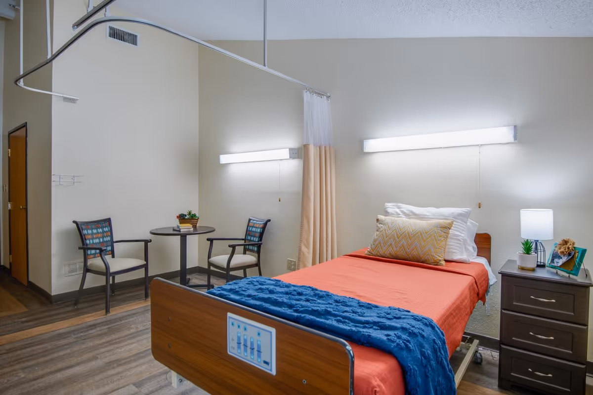 A well-lit room in a healthcare facility featuring a single hospital bed with orange bedding and a blue blanket. The bed has a control panel on the footboard. Next to the bed is a nightstand with a lamp, a small plant, and a framed photo. On the left side of the room, there is a small round table with two chairs. A privacy curtain is partially drawn around the bed. The floor is wood-patterned, and the walls are light-colored.