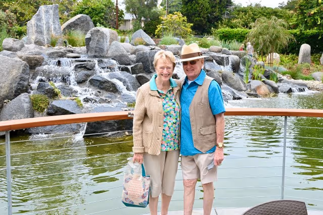 Two adults standing on a deck in front of a pond with a rock waterfall and landscaped gardens.
