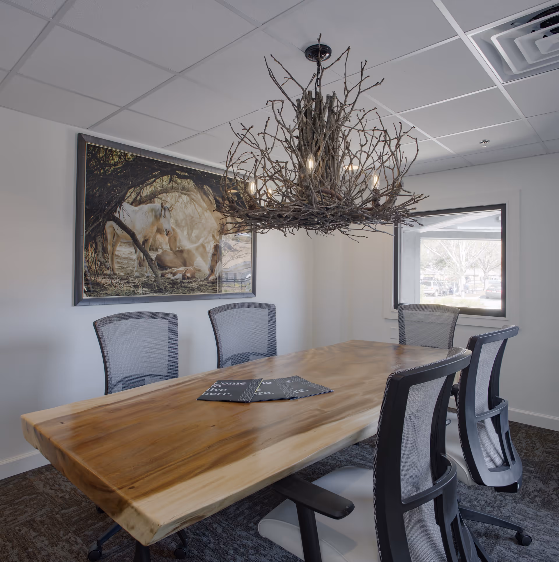 A modern conference room with a large wooden table surrounded by five mesh office chairs. A unique chandelier made of intertwined branches hangs above the table. On the wall, there is a framed picture of horses under a tree. A window lets in natural light, showing a view of the outside parking area.