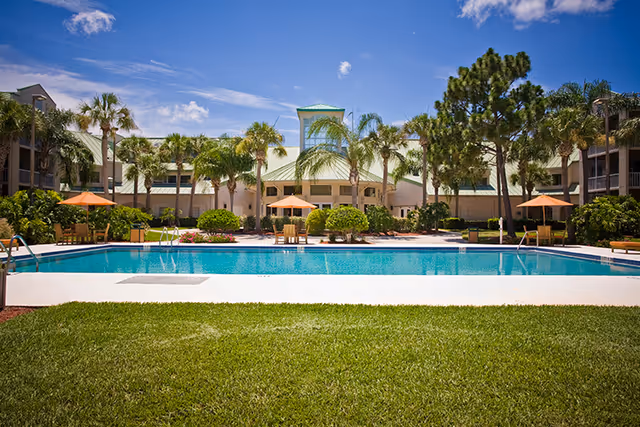 Rectangular outdoor swimming pool with lounge chairs, umbrellas, and palm trees in front of a low-rise building under a blue sky.