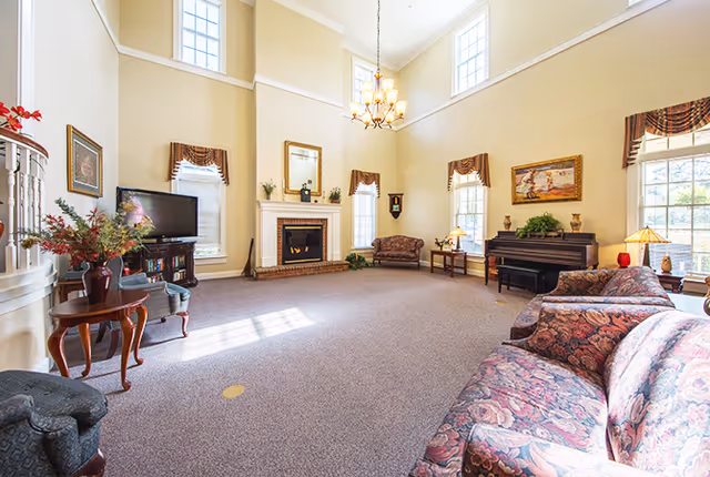 A spacious living room with high ceilings and large windows letting in natural light. The room features a fireplace with a mirror above it, a television on a stand, multiple upholstered chairs and sofas with floral patterns, a wooden piano, side tables with lamps, and framed artwork on the walls. The carpet is light brown, and the walls are painted a soft beige color.