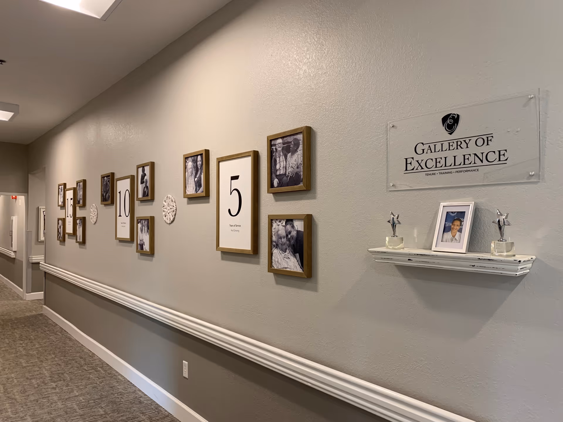 A hallway with a gallery wall featuring framed photos and numbers representing years of service. There is a sign that reads 'Gallery of Excellence' with the words 'Tenure - Training - Performance' underneath. Below the sign is a small shelf holding a framed photo and two star-shaped trophies.