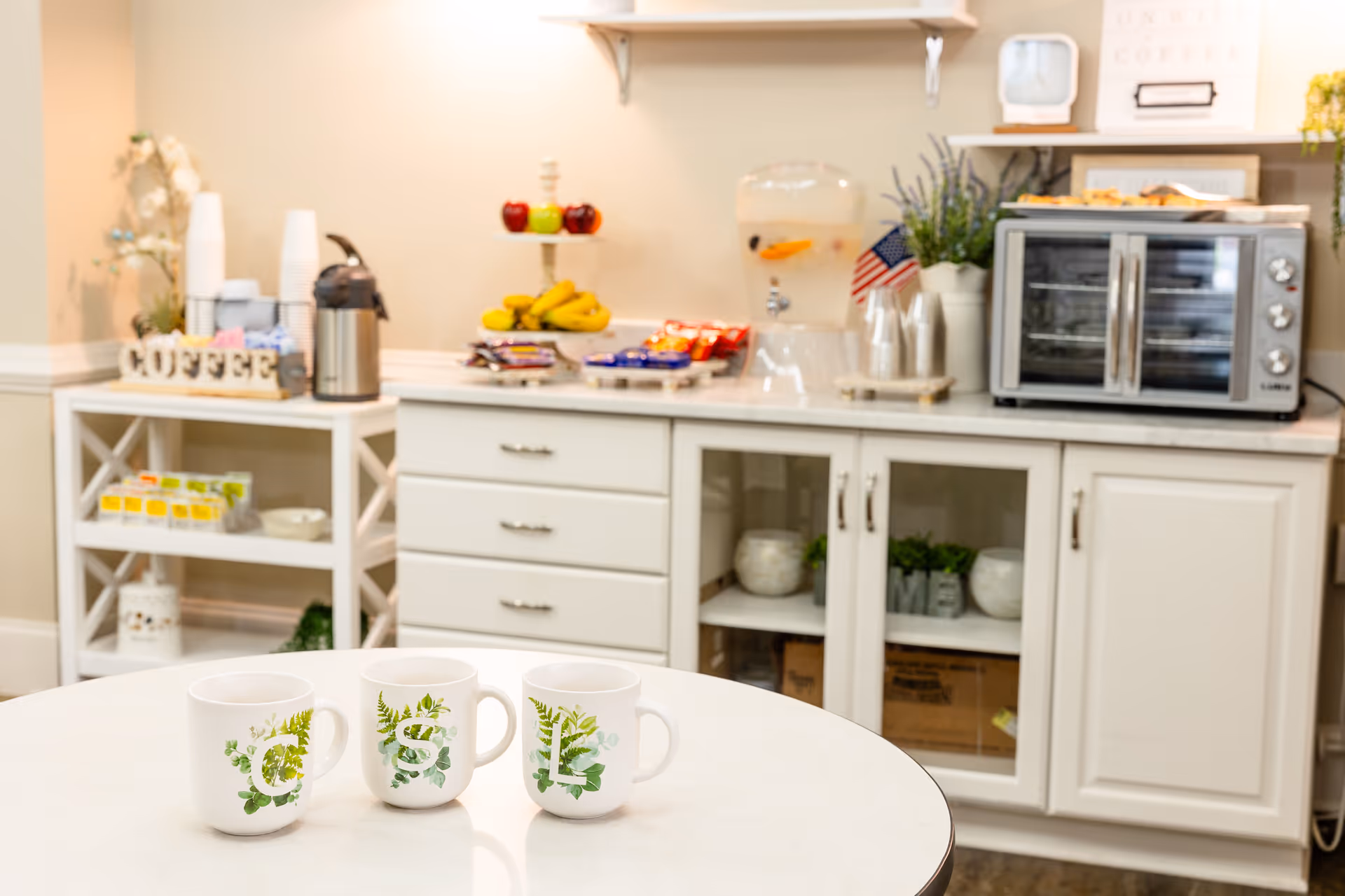 A small kitchenette/coffee station with three decorative mugs on a round table in the foreground and cabinets, a coffee urn, fruit, a water dispenser, and a toaster oven on the counter behind.