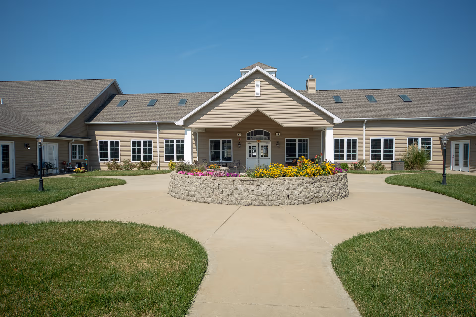 Exterior view of a single-story senior living facility building with beige siding, multiple windows, and a central covered entrance. In front of the entrance is a circular stone planter filled with colorful flowers, surrounded by a paved walkway and green grass. The sky is clear and blue.