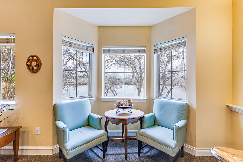 Two light-blue upholstered armchairs flank a small round wooden table in a bay window overlooking a lake.