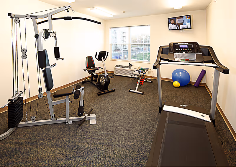 A small fitness room with exercise equipment including a multi-gym machine, a stationary bike, a treadmill, an exercise ball, a yoga mat, and a small set of dumbbells. A window lets in natural light, and a TV is mounted on the wall.