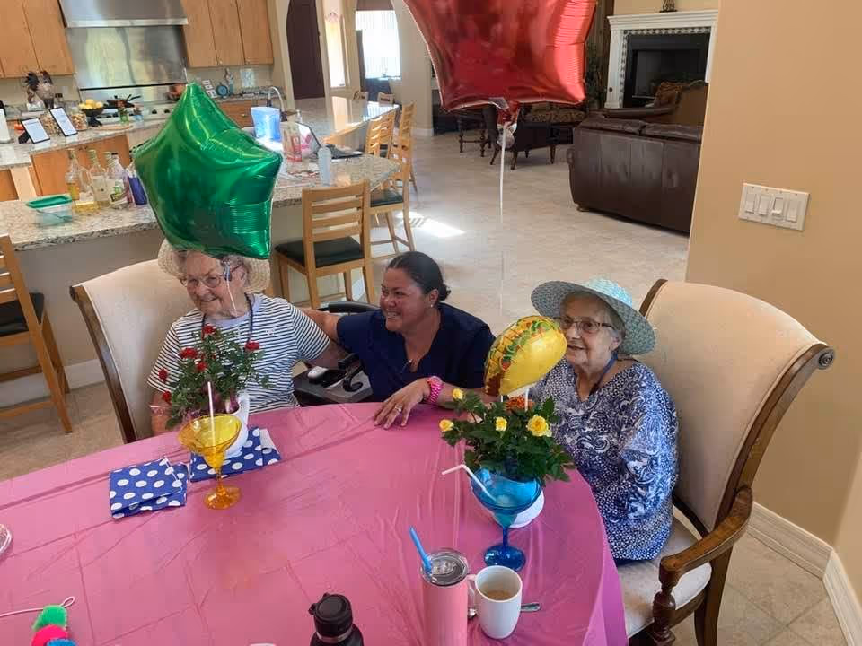 Three women sitting around a pink-covered table with balloons and flower centerpieces in a senior living common dining area.