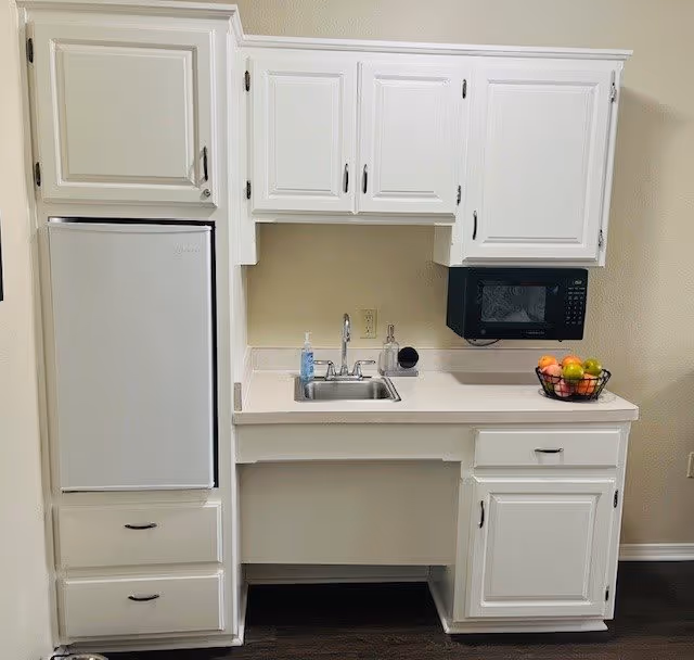 A compact kitchen area with white cabinets, a small refrigerator, a sink with a faucet, a black microwave mounted above the counter, and a fruit basket containing apples and oranges on the countertop.