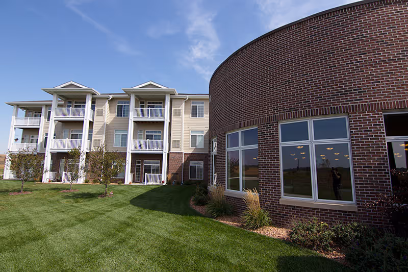 Exterior of a senior living facility showing a curved brick common area next to a three-story building with balconies and a manicured lawn.