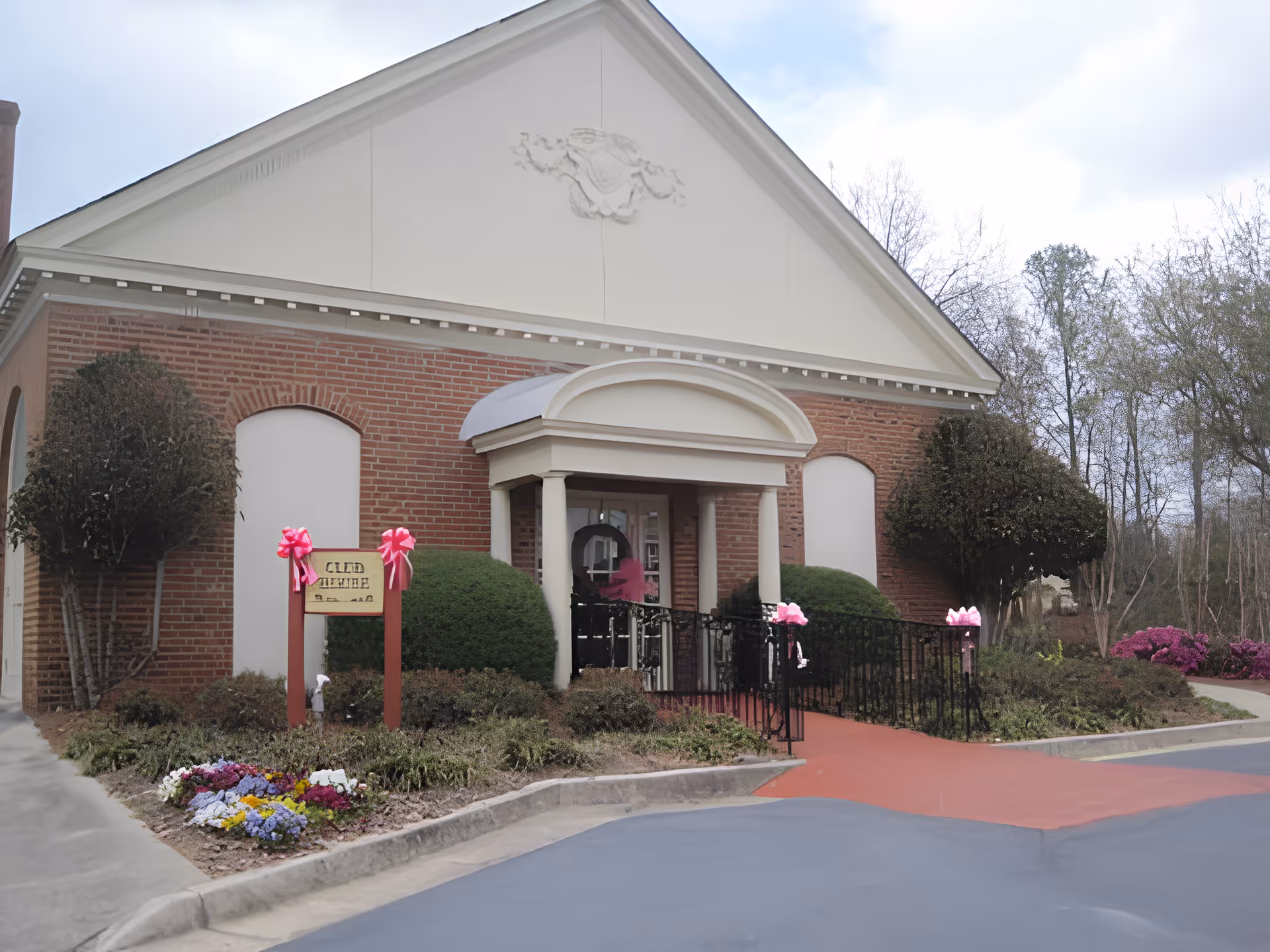 Exterior view of a brick building with a white triangular pediment and a small covered entrance supported by columns. The entrance has a ramp with black railings decorated with pink bows. There are trimmed bushes and flower beds with colorful flowers in front of the building. Trees and a cloudy sky are visible in the background.