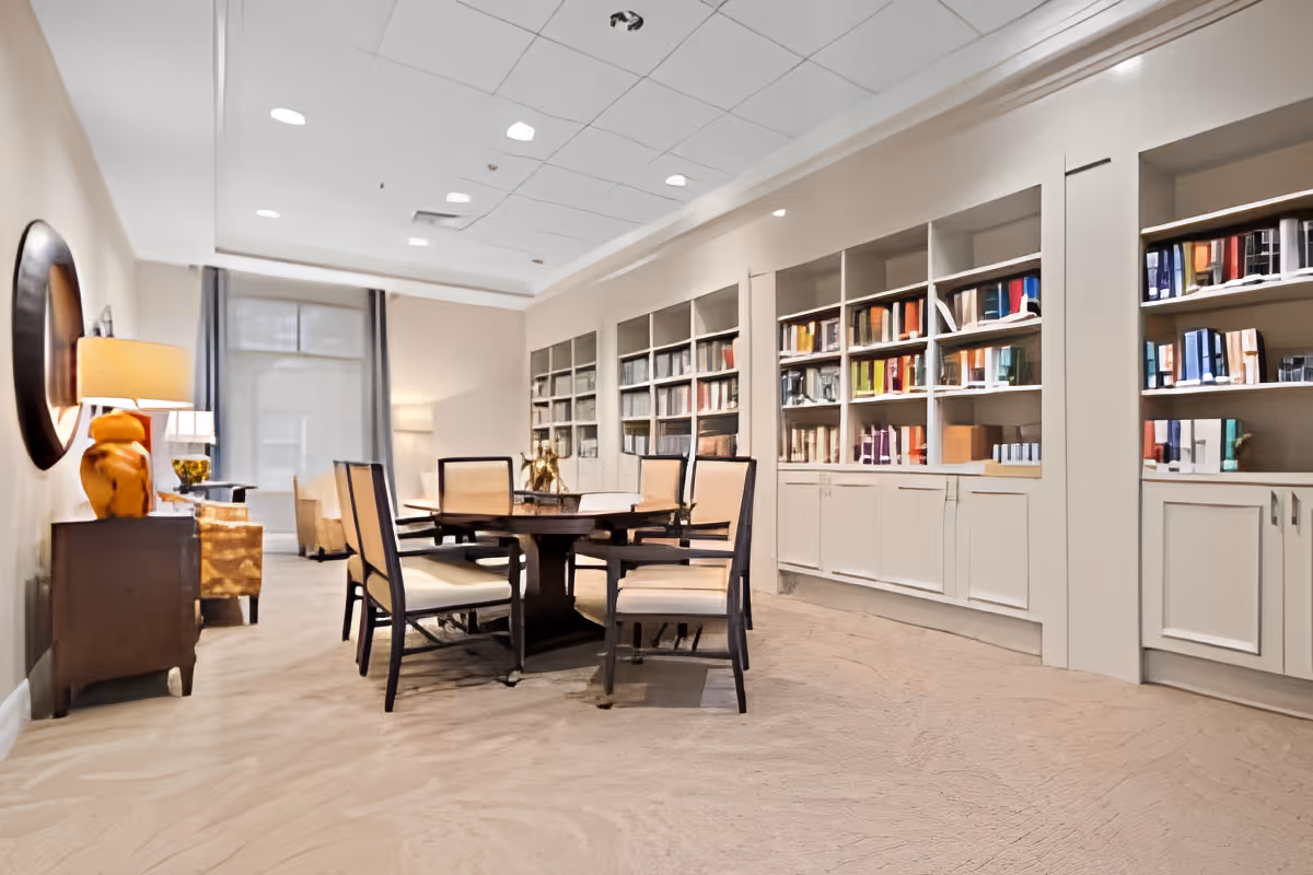 A bright communal library-style lounge with a central round table and chairs and built-in bookshelves along the wall.