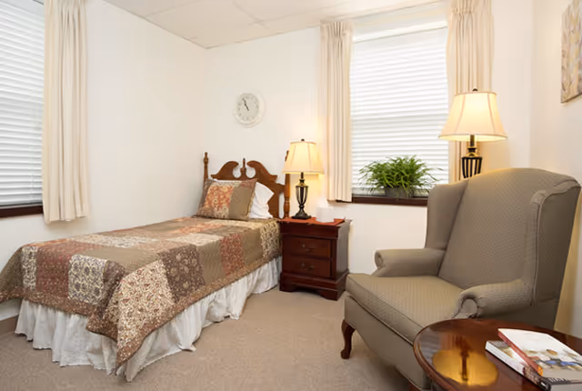 A cozy bedroom with a single bed covered in a patchwork quilt, a wooden nightstand with a lamp and tissue box, a comfortable armchair, a small round table with books, two windows with blinds and curtains, and a wall clock.
