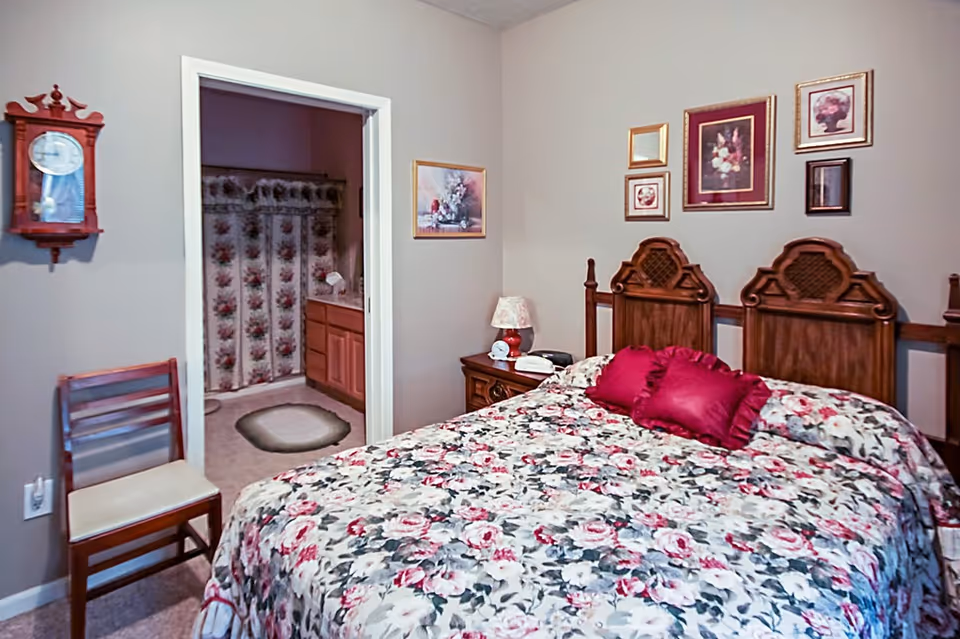 A bedroom with a floral bedspread on a wooden bed frame, two red pillows, a wooden nightstand with a lamp and telephone, a wooden chair with a cushion, and floral artwork on the walls. An open doorway reveals a bathroom with a floral shower curtain and a rug on the floor.