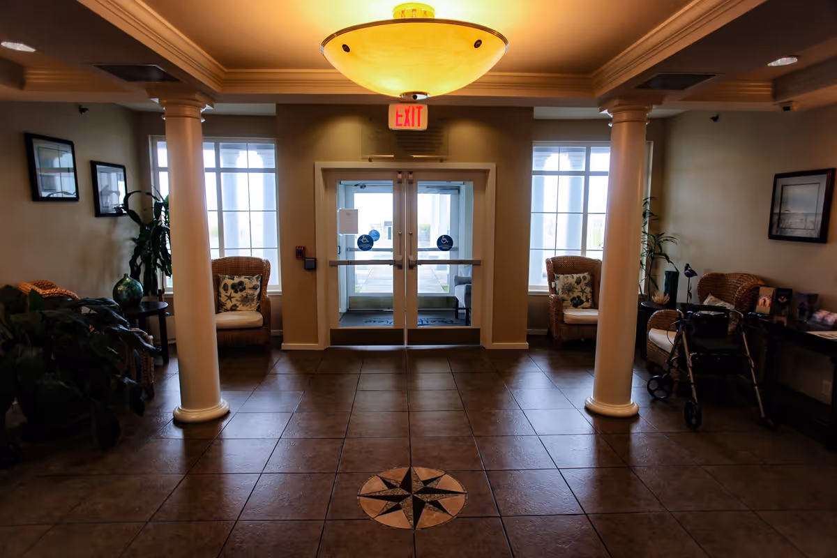 Interior view of a senior living community lobby with tiled floor featuring a compass design, two white columns, wicker chairs with cushions, plants, framed pictures on the walls, a walker, and double glass exit doors with windows on either side.