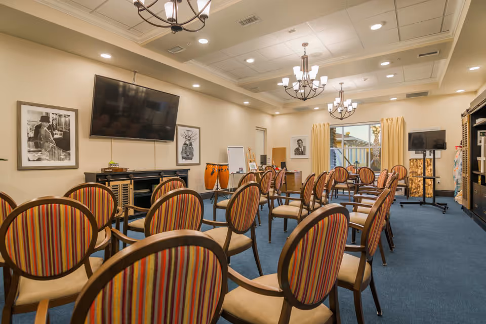 A well-lit meeting or activity room with rows of wooden chairs featuring striped upholstery arranged facing a large flat-screen TV mounted on the wall. The room has blue carpet, beige walls, two chandeliers, framed black and white photographs, a whiteboard, conga drums, and a window with beige curtains.