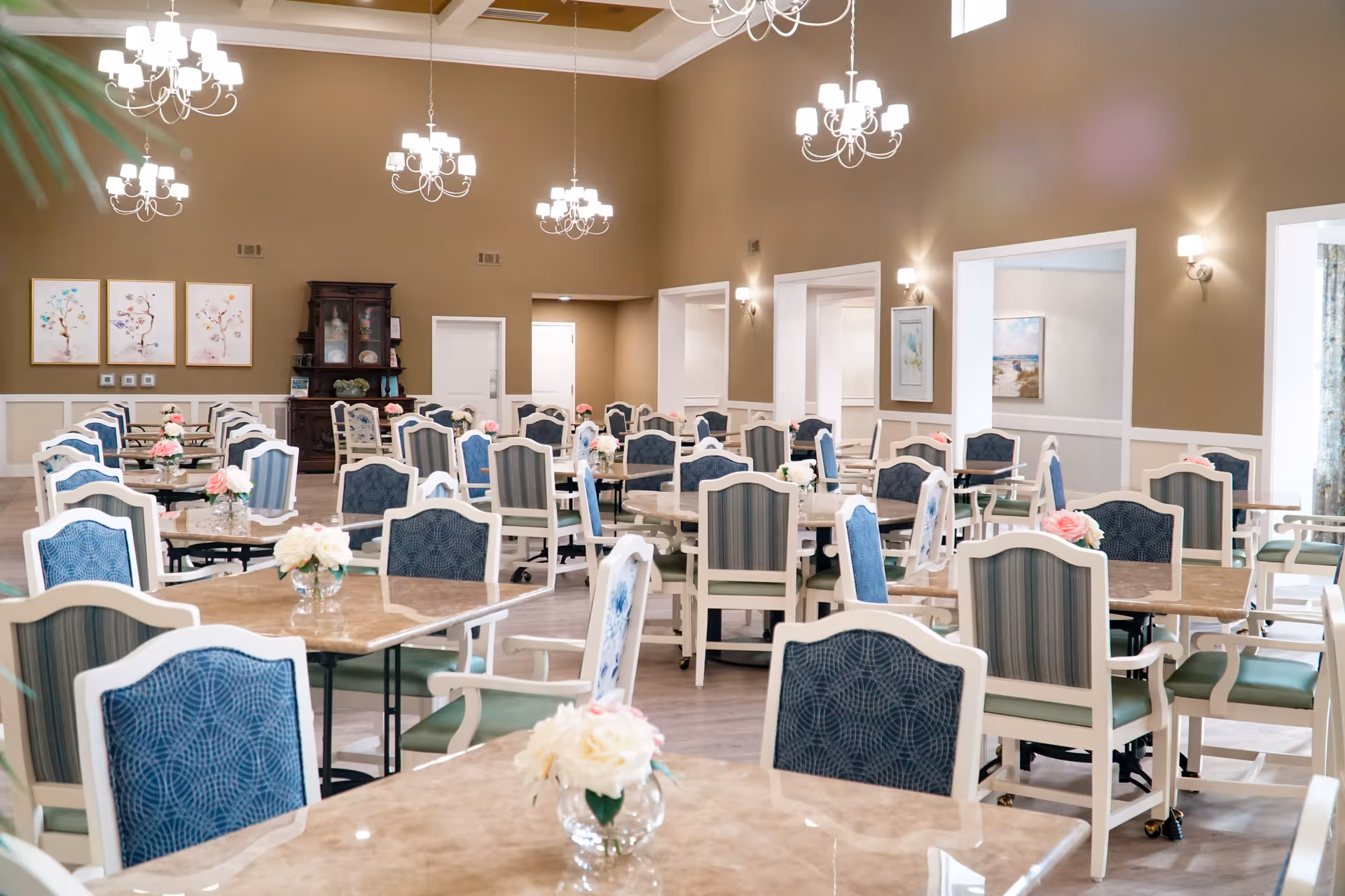A spacious dining room with multiple tables and chairs arranged neatly. Each table has a small vase with flowers. The room features beige walls, white trim, several chandeliers hanging from the ceiling, and framed artwork on the walls.
