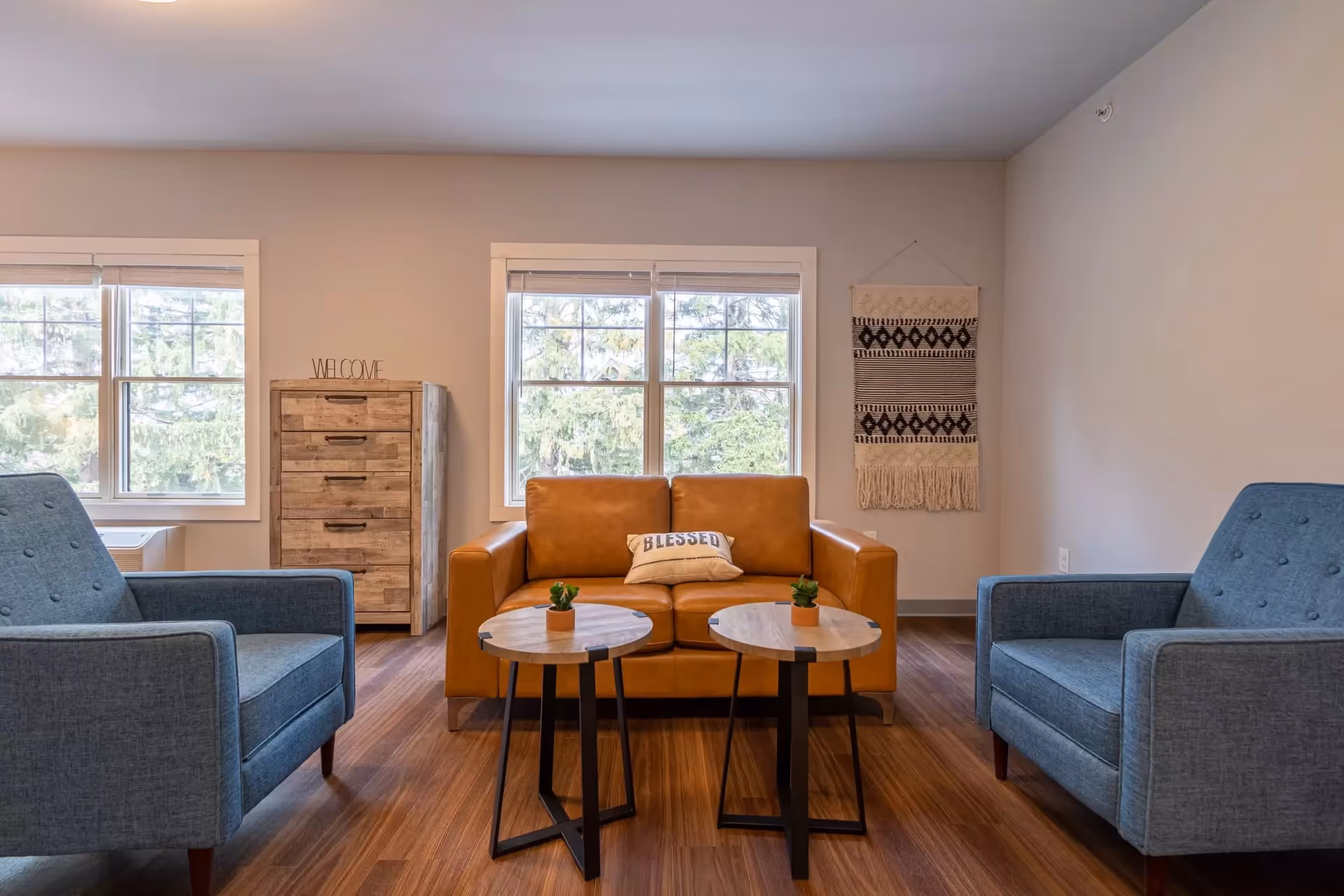 A cozy living room with a brown leather loveseat featuring a 'BLESSED' pillow, two blue armchairs, two round wooden side tables with small potted plants, a wooden chest of drawers with a 'WELCOME' sign on top, and a decorative woven wall hanging. Large windows let in natural light and show greenery outside.