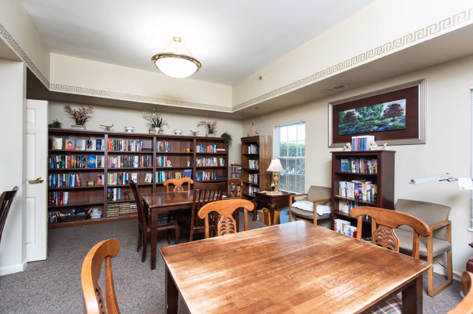 A cozy reading room with wooden tables and chairs, bookshelves filled with books, decorative plants on top of the shelves, a window letting in natural light, and a framed painting on the wall.