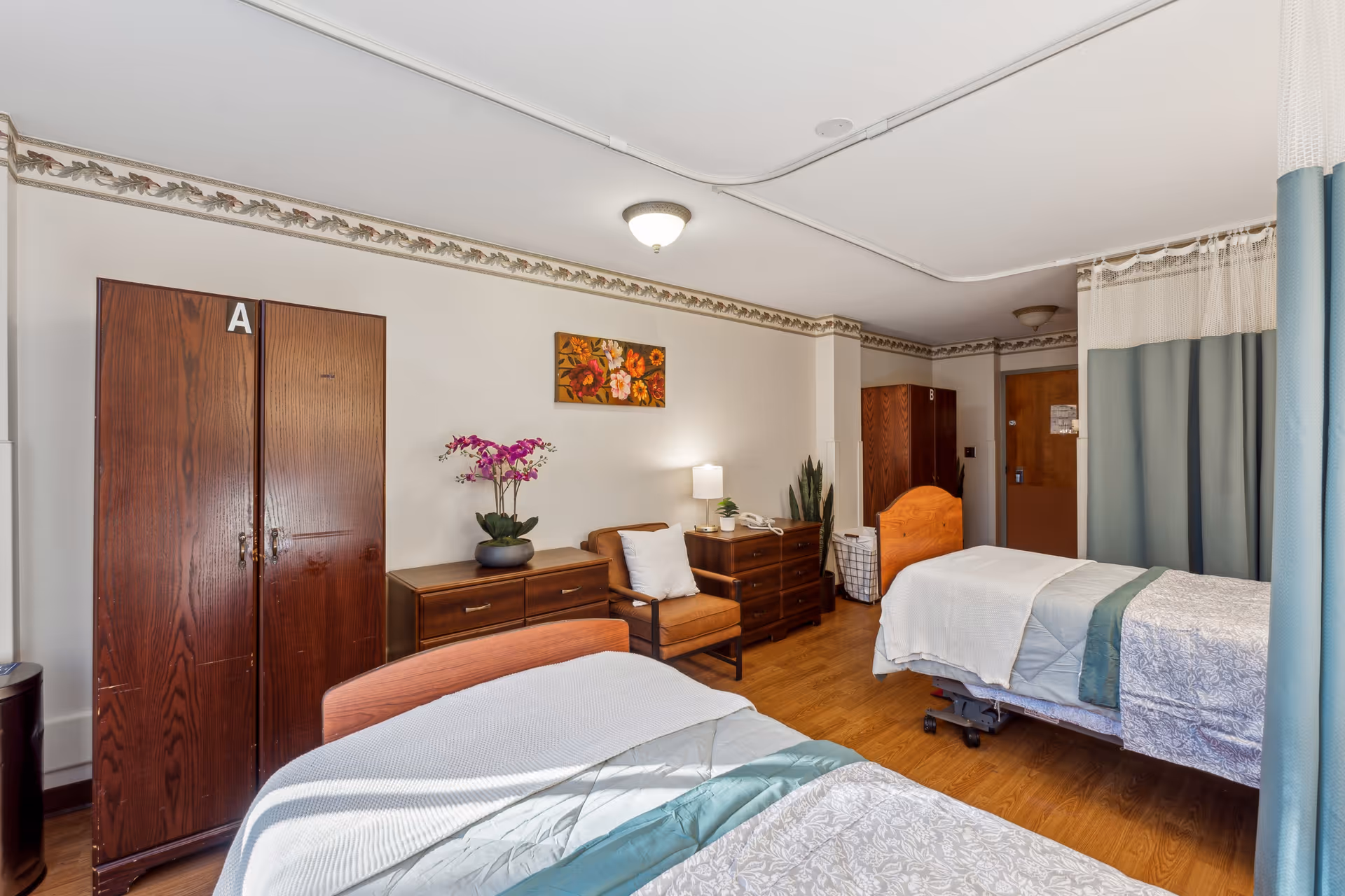 A nursing facility room with two beds separated by a curtain. Each bed has white and teal bedding. The room features wooden furniture including two wardrobes labeled A and B, a dresser, a chair with a cushion, and a small table with a lamp and a potted plant. A floral painting hangs on the wall above the chair. The floor is wooden, and the room has a ceiling light fixture.