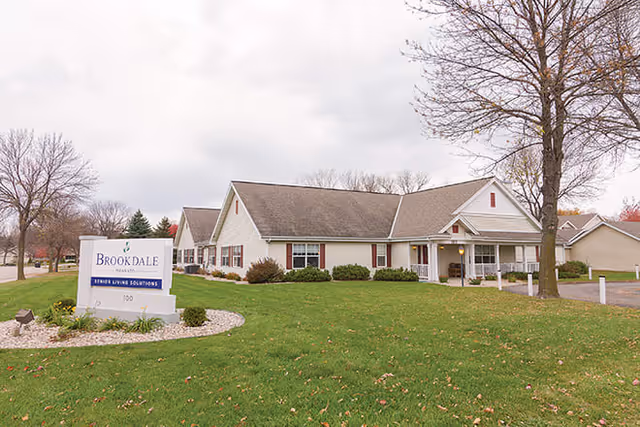 Exterior view of Brookdale Mankato senior living facility showing a single-story building with a pitched roof, surrounded by a green lawn and trees with some leaves fallen. A white sign in the foreground reads 'Brookdale Mankato Senior Living Solutions'.