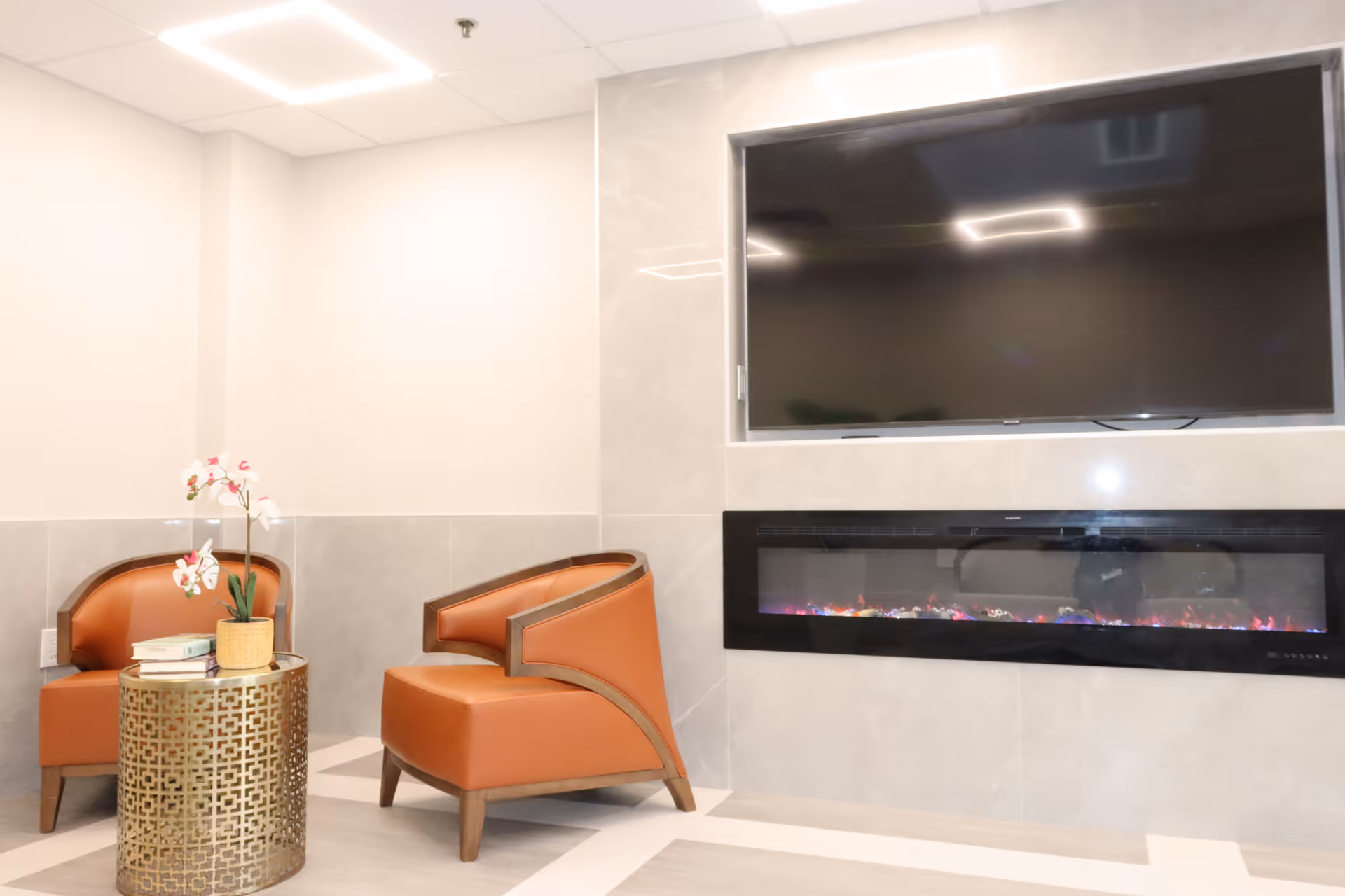 A cozy seating area with two brown leather chairs and a round gold metal side table with a potted orchid and books on it. Mounted on the wall above a modern electric fireplace is a large flat-screen TV. The room has light-colored walls and tiled flooring.