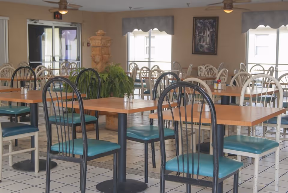A dining room with multiple wooden tables and chairs with teal cushions. The room has tiled floors, large windows with valances, a ceiling fan, and a decorative plant in the center. There are salt and pepper shakers on the tables and a framed picture on the wall.